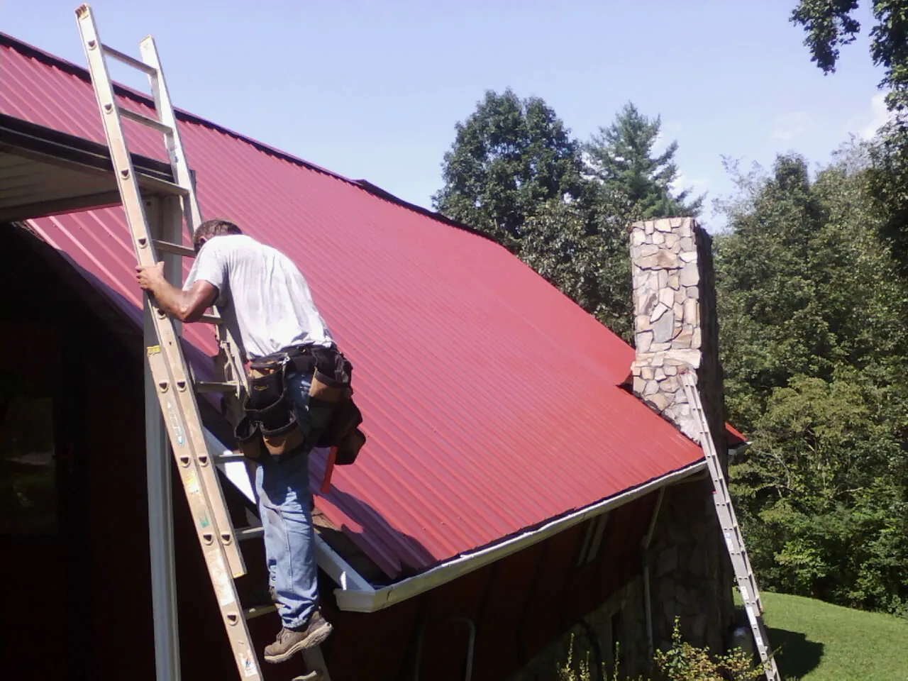 Roofer on a ladder beside a red metal roof and stone chimney — Soffit & Fascia Repair by Roofing Brothers in Appling